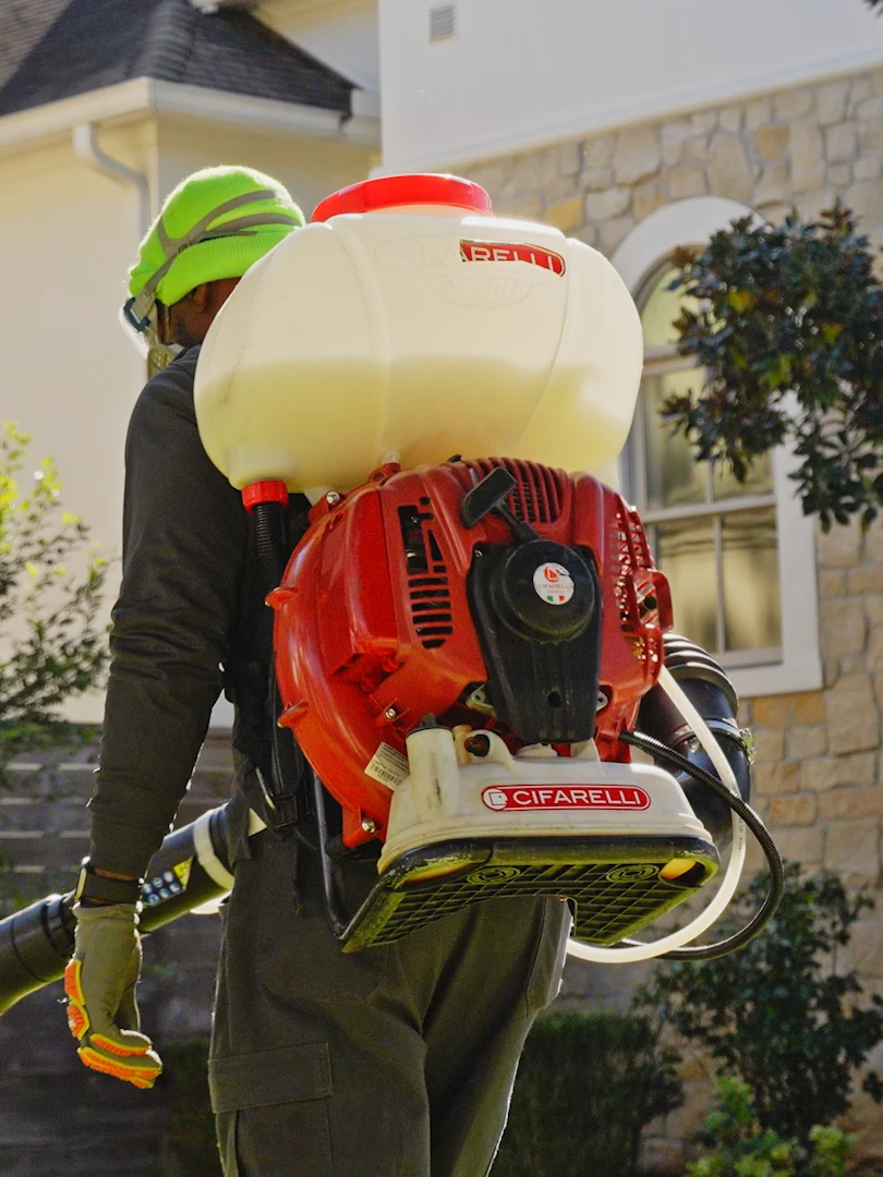 Pest control technician in protective gear carries a backpack mist blower toward the landscaping of a stone-and-stucco house.